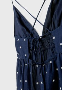 Granatowa sukienka z głębokim dekoltem w kształcie litery V, krzyżującymi się ramiączkami i elastycznym pasem. Posiada białe hafty w kształcie gwiazdek rozproszone na materiale.