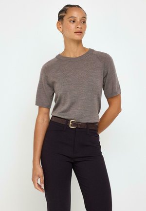 Kvinde med fletninger iført en kortærmet brun sweater, sorte bukser og et tekstureret mørkebrunt bælte mod en ensartet lys baggrund.