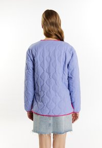 Quiltet lilla jakke med pink detaljer, der har et tekstureret bølgemønster. Bæres over en denimnederdel med frynset kant. Set fra ryggen.
