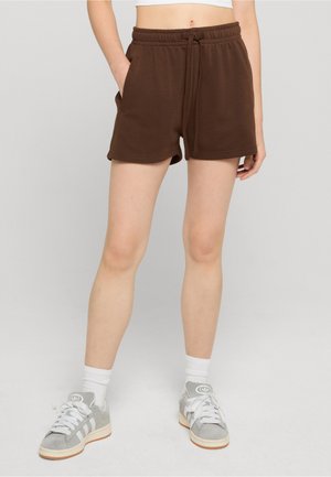 Person iført brune shorts med snøre, hvide sokker og grå sneakers, stående med den ene hånd i lommen mod en ensfarvet baggrund.
