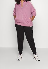 Sudadera con capucha lila claro con cremallera, rayas blancas a los lados y bolsillos tipo canguro, combinada con pantalones jogger negros. La modelo lleva calzado blanco con detalles.