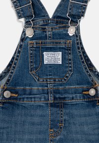 Tmavě modré denimové kalhoty na kšandy s kovovými stříbrnými prvky, nastavitelnými popruhy a přední kapsou s nášivkou. Viditelné kontrastní švy.