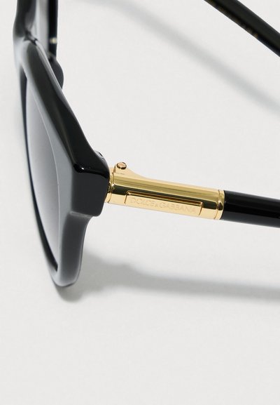 Juodi akiniai nuo saulės su kvadratiniais rėmeliais, auksiniu akcentu vyriai, blizgiu paviršiumi ir tamsiais lęšiais. Ant metalinių detalių išgraviruotas prekės ženklo logotipas.