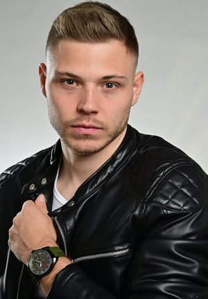 Das Modell trägt eine schwarze, gesteppte Lederjacke mit Reißverschlussdetails, ein weißes Hemd darunter und eine runde grüne Uhr mit schwarzen Akzenten.