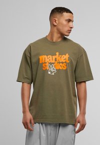 Olivenfarget oversized t-skjorte laget av bomull. Har oransje fet tekst "market studios" og et fredssymbol grafikk. Korte ermer.