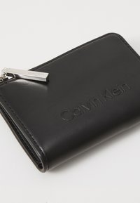 Portafoglio in pelle nera con logo "Calvin Klein" in rilievo, texture liscia e una cerniera in metallo argentato. Design rettangolare compatto.