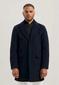 Marinewollen jas met een notch-kraag, twee voorzakken en een rechte snit. G gedragen over een zwart ritssluiting kledingstuk met een gestructureerde afwerking.