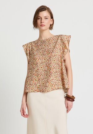 Frau trägt eine ärmellose Bluse mit floralem Muster und Rüschenärmeln sowie einen beigen Rock, ergänzt durch braune Armreifen.