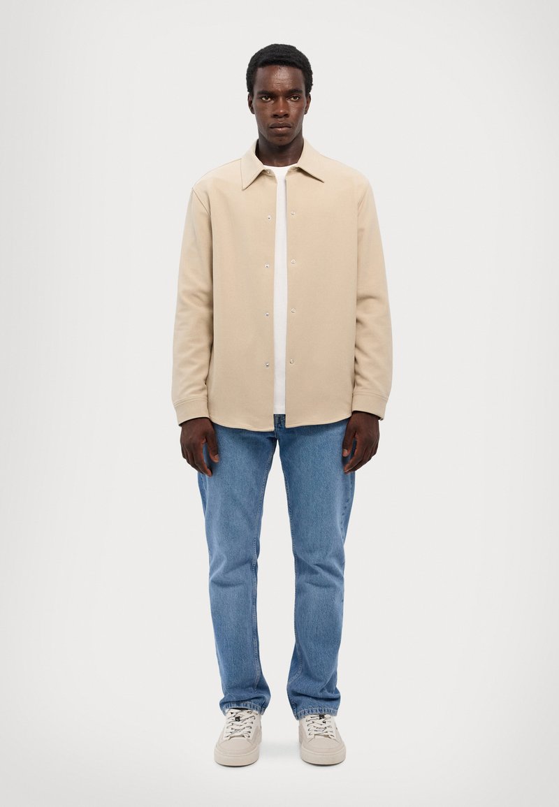 Beige button-up shirt gemaakt van zachte stof met een puntige kraag, gecombineerd met lichtblauwe jeans en witte sneakers. Eenvoudig ontwerp en schone lijnen.