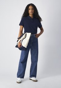 Polo shirt marine, jean bleu taille haute et baskets blanches. Le modèle tient un grand sac fourre-tout crème et marine avec deux anses.