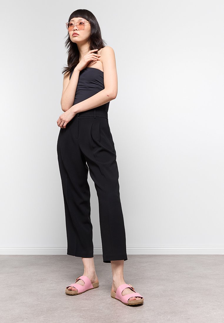 Femme portant un haut sans bretelles noir et un pantalon, des sandales à glisser roses et des lunettes de soleil, se tenant contre un fond clair.
