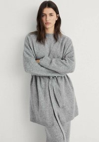 Cardigan grigio lavorato a maglia, stile avvolgente, con maniche ampie e chiusura a fiocco. Texture morbida e lunghezza fino a metà coscia, con dettagli a coste sull'orlo.
