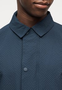 Κοντινό πλάνο ενός ατόμου που φοράει υφασμάτινο σκούρο μπλε πουκάμισο με γιακά και ορατά κουμπιά, με έμφαση στον λαιμό και το πάνω μέρος του στήθους.