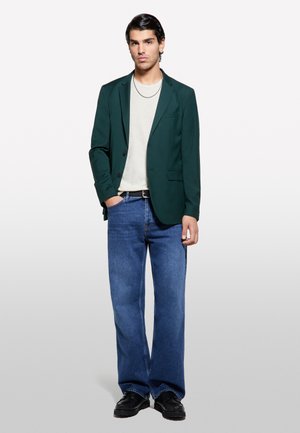 Blazer verde scuro sopra una camicia grigia chiara, abbinato a jeans blu a gamba larga e scarpe nere. Una collana d'argento evidenzia il look.