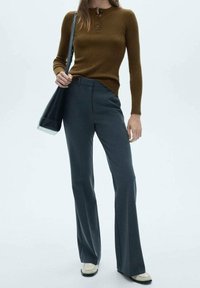 Top long à manches longues marron à côtes avec une patte de boutonnage, associé à un pantalon évasé gris foncé et des chaussures couleur crème, tenant un sac noir.