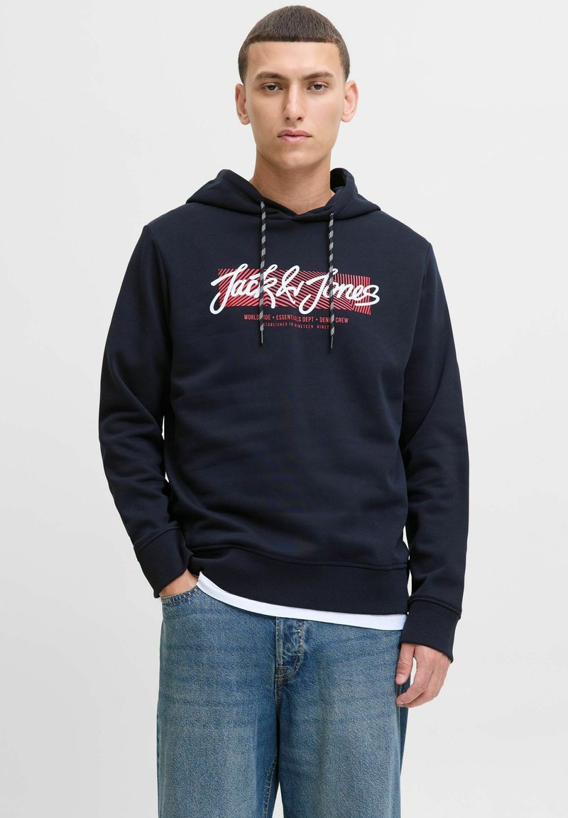 Granatowy hoodie z białym i czerwonym nadrukiem logo. Posiada kaptur ze sznurkiem i ściągacze przy mankietach, zestawiony z jasnoniebieskimi dżinsami.