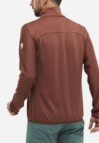 Brown zip-up jacket with a textured pattern, high collar, and logo on upper left sleeve. Worn over light green pants.