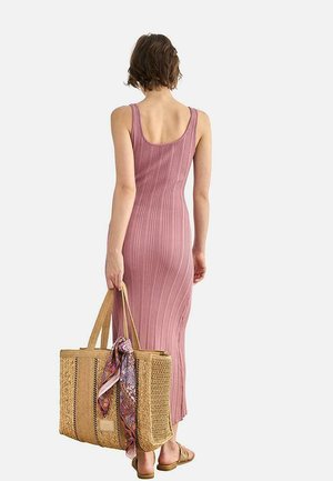 Mujer con un vestido sin mangas de canalé rosa, sosteniendo un bolso grande de tela trenzada con un pañuelo colorido anudado al asa, de pie sobre un fondo blanco.