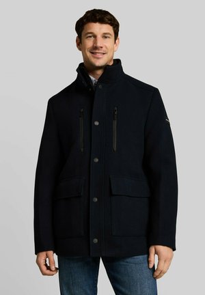 Uomo che indossa un cappotto nero con bottoni, tasche sul petto con cerniera e tasche frontali con patta, in piedi davanti a uno sfondo grigio chiaro uniforme.