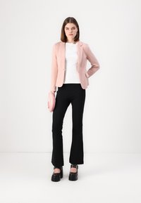 Svetle růžový tailored blejzr, bílý top, černé rozšířené kalhoty a robustní černé platformové boty. Drží zaoblenou růžovou psaníčko.