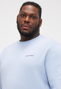 Világoskék, pamut keverékből készült pulóver. Kerek nyakkivágással és hímzett "Calvin Klein" logóval rendelkezik a mellkason. Simább textúrájú.