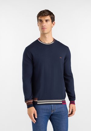 Giovane uomo che indossa un maglione blu navy con polsini e orlo a coste a righe, abbinato a jeans blu, in piedi davanti a uno sfondo bianco.