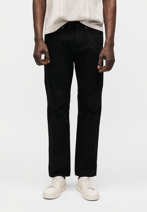 Homme portant un pantalon noir droit, un haut texturé écru à manches courtes et des baskets couleur crème, debout sur un fond uni.