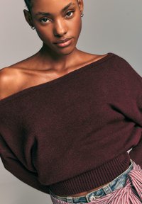 Burgundska pletena pulover s off-shoulder izrezom in rebrastim robom, v kombinaciji z modrimi denim kratkimi hlačami, ki imajo na pasu povezan črtast pas.