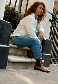 Bottes courtes en cuir marron avec des panneaux latéraux élastiques, un bout arrondi et un petit talon carré, portées avec un jean bleu clair et un pull blanc.