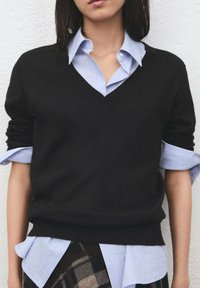 Čierny pulóver s výstrihom do V cez svetlomodrú prúžkovanú košeľu, s vyhrnutými rukávmi a vzorovanou sukňou pod tým.