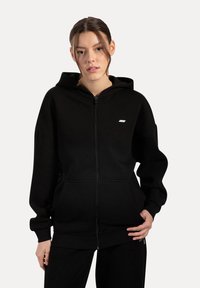 Czarna bluza z kapturem na zamek, wykonana z miękkiego materiału, z przednią kieszenią, ściągaczami na rękawach i małym logo na lewej piersi. Prosty design.