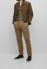 Brun lättviktsjacka med svarta detaljer, brun texturerad tröja och beige byxor, tillsammans med svarta sneakers.