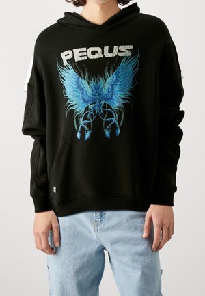 Henkilö, jolla on päällään musta huppari, jossa suuri sininen abstrakti siivekäs olento ja valkoinen "PEQUS"-teksti, yhdistettynä vaaleansinisiin farkkuihin.