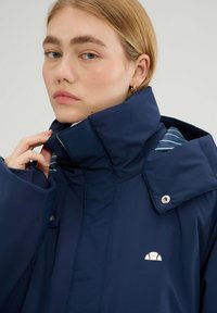 Granatowa wodoodporna kurtka z wysokim kołnierzem, napami i akcentami na ramionach; posiada paskowany materiał wewnętrzny oraz logo na piersi.