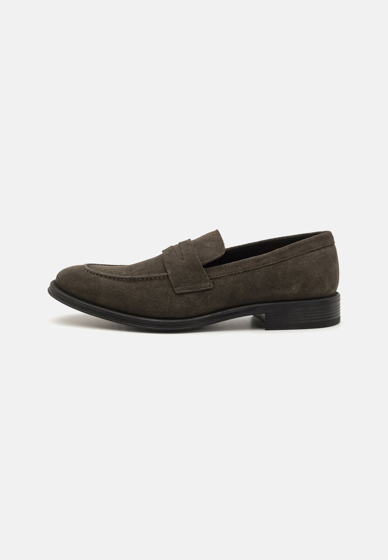 Mocasines de ante marrón con un bajo tacón negro, que presentan una textura suave y una amplia correa horizontal en la parte delantera.