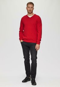 Rode v-halstrui van zachte stof, gecombineerd met zwarte slim-fit jeans en zwarte veterschoenen, staande tegen een neutrale achtergrond.