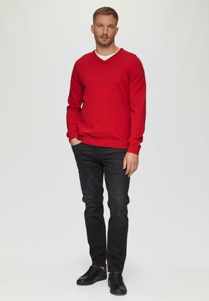 Roter V-Ausschnitt-Pullover aus weichem Stoff, kombiniert mit schwarzen, engen Jeans und schwarzen Schnürschuhen, vor einem neutralen Hintergrund stehend.