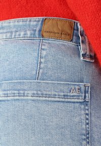 Heledad sinine teksapüksid, mille vööl on nahkplekk, millel on silt "American Eagle", tagaküljel on tasku ja peened õmblusdetailid.