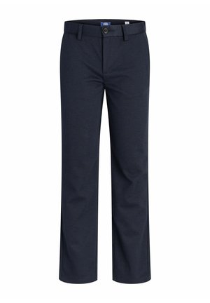 Ragazzo in piedi che indossa una camicia azzurro chiaro con bottoni, pantaloni scuri e scarpe da ginnastica bianche su uno sfondo bianco uniforme.