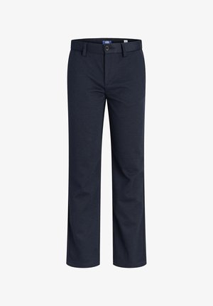 Junge steht, trägt ein hellblaues Hemd mit Knöpfen, dunkle Hose und weiße Turnschuhe vor einfachem weißem Hintergrund.