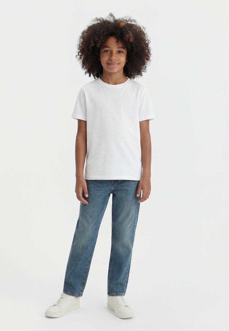 Kind mit lockigem Haar steht frontal, trägt ein schlichtes weißes T-Shirt, blaue Jeans und weiße Turnschuhe vor einem weißen Hintergrund.