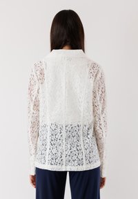 Blusa bianca in pizzo con colletto, maniche lunghe e un motivo floreale traslucido. La parte posteriore ha un taglio leggermente più lungo con cuciture verticali.