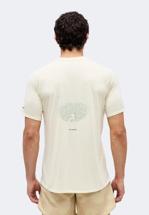 Άνδρας με σγουρά σκούρα μαλλιά που φορά ανοιχτό μπεζ ελαφρύ κοντομάνικο μπλουζάκι για τρέξιμο και ταιριαστά σορτς, φαίνεται από πίσω.