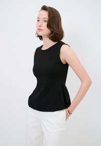 Schwarzes Peplum-Top aus strukturiertem Stoff, mit rundem Kragen und ärmellosem Design. Kombiniert mit weißen Hosen aus glattem Material.