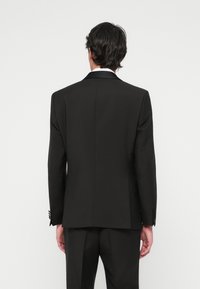 Homme aux cheveux foncés portant un costume noir et une chemise blanche, se tenant de dos contre un fond blanc uni.