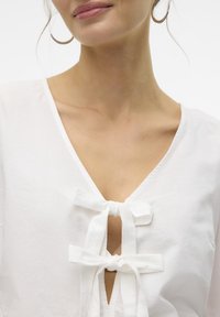 Blusa bianca in cotone con scollatura a V e due dettagli a fiocco sulla scollatura. Tessuto morbido; design minimalista con linee pulite.