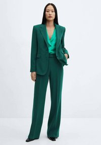 Groene pakset bestaande uit een diep groene blazer, bijpassende wijde broek en een lichtgroene blouse. De stof lijkt glad met een getailleerde pasvorm.