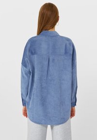 Blaues, langärmliges Hemd aus weichem, strukturiertem Stoff. Verfügt über einen Kragen, einen Faltenwurf auf der Rückseite und einen abgerundeten Saum.