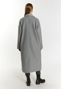 Grijze wollen jas met een middellange, brede kraag en lange mouwen. Gecombineerd met zwarte enkellaarzen, gezien van achteren. Eenvoudig design.
