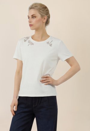 Vaaleahiuksinen nainen, jolla on hiukset nutturalla, pukeutuneena valkoiseen, lyhythihaiseen toppiin, jossa on koristeellisia hopeisia somisteita olkapäillä, ja tummiin housuihin.